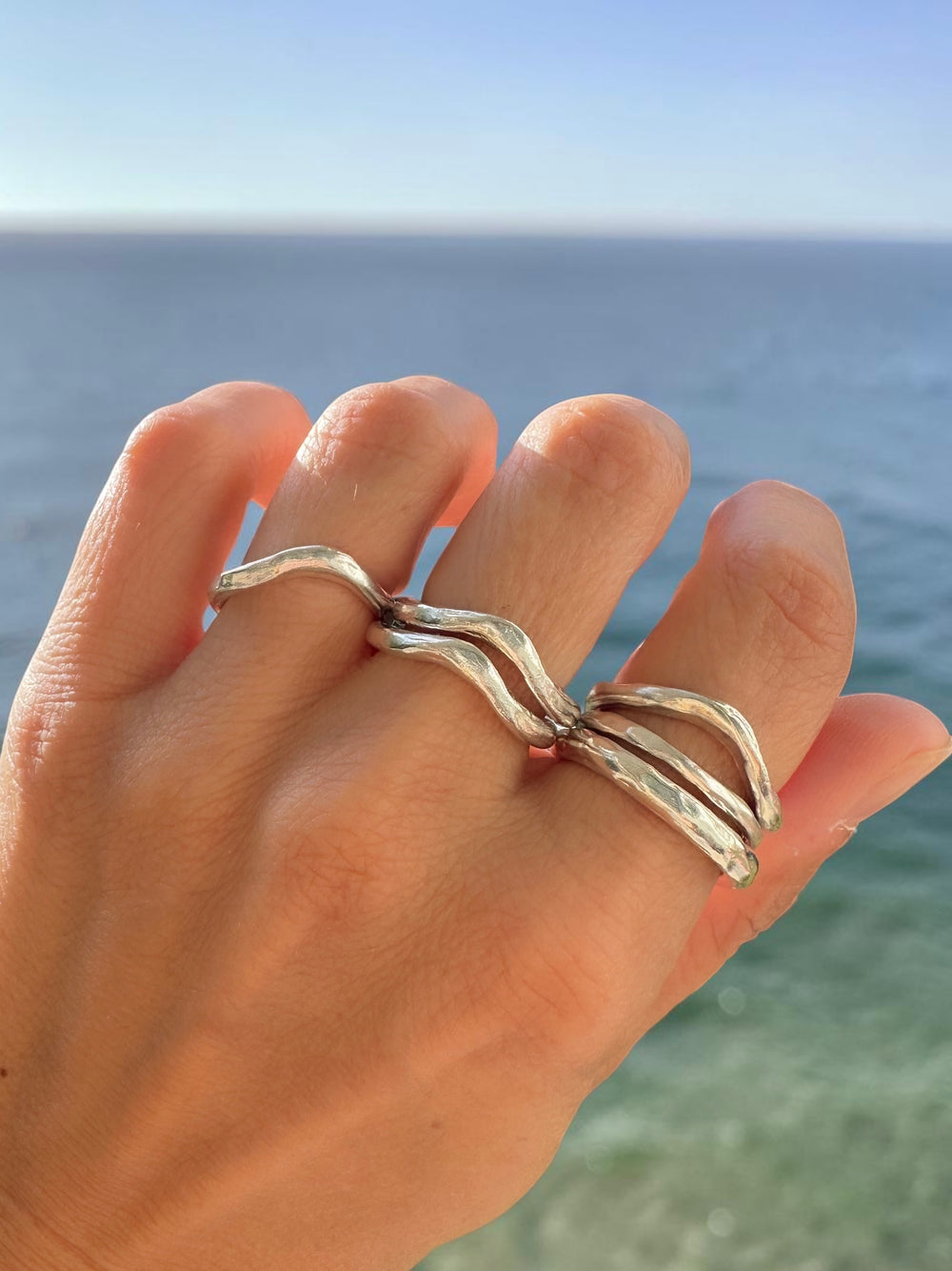 Sterling silver wave stacking rings worn across multiple fingers against a clear blue ocean, featuring an organic, coastal-inspired design. 