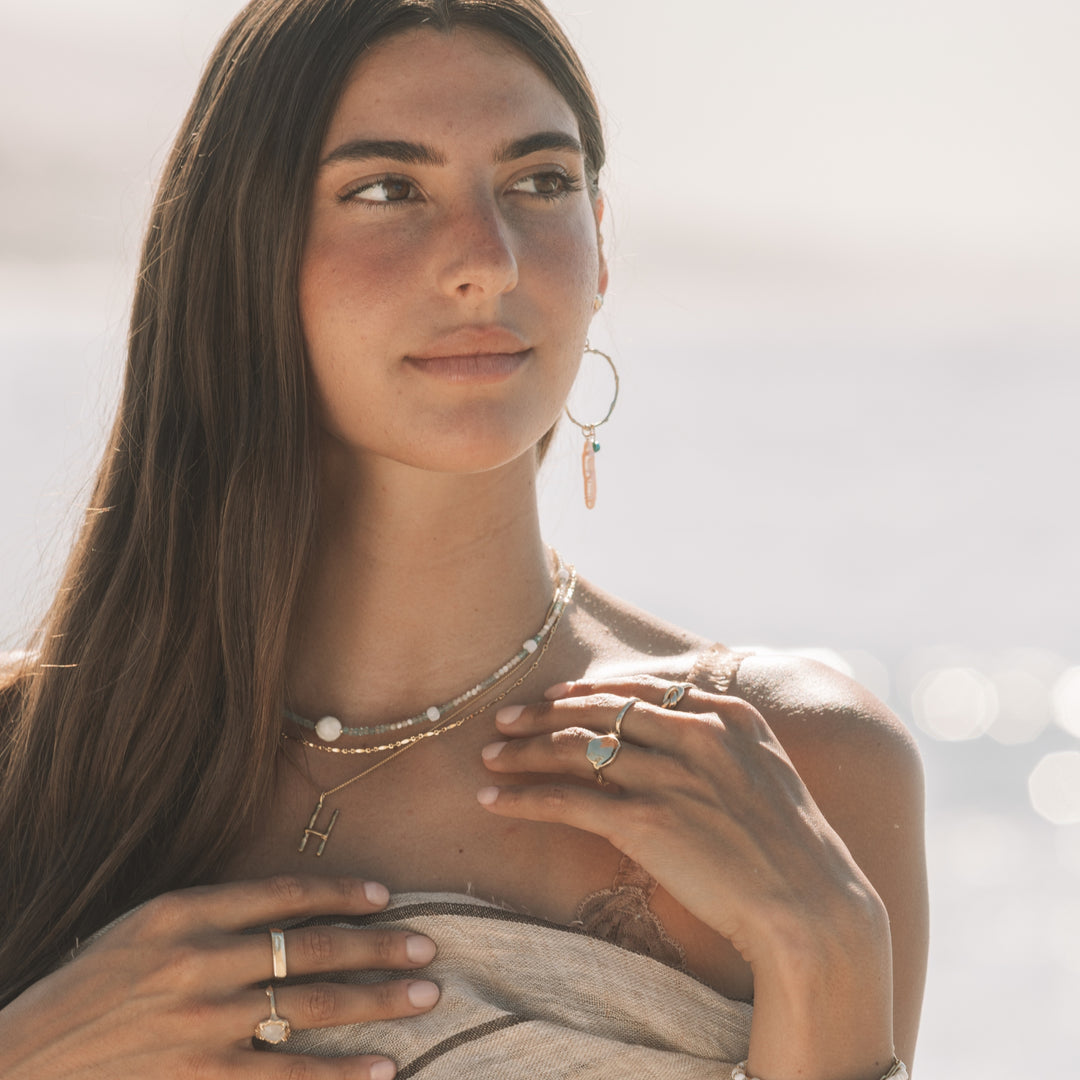 Woman wearing Seaside Hoops with turquoise and pearl drops, layered gold necklaces, gold rings and linen wrap by the ocean, handmade coastal boho jewelry by Blooming Lotus Jewelry.