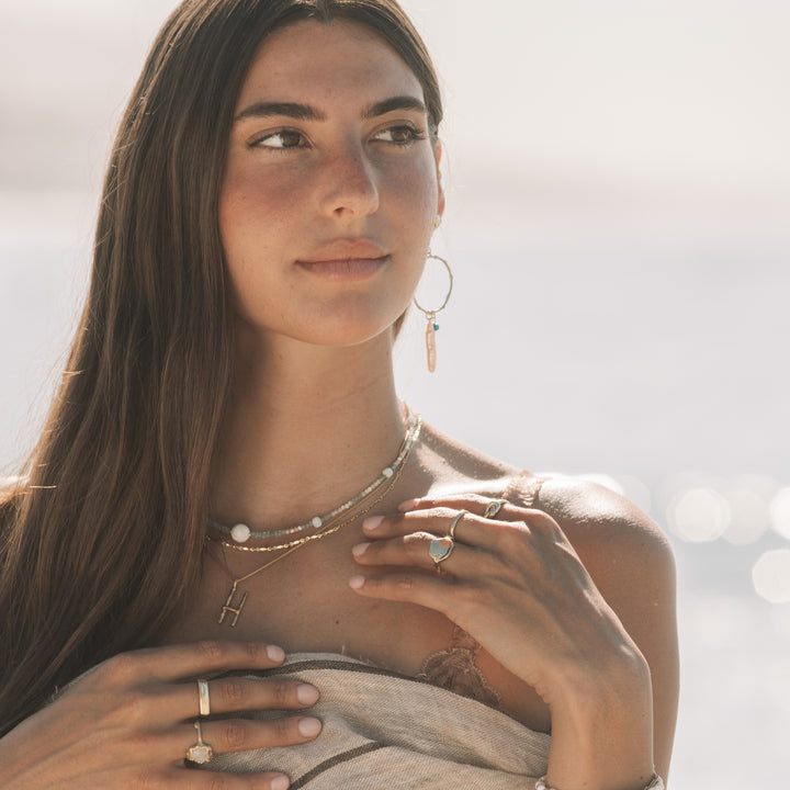 Woman wearing Seaside Hoops with turquoise and pearl drops, layered gold necklaces, gold rings and linen wrap by the ocean, handmade coastal boho jewelry by Blooming Lotus Jewelry.