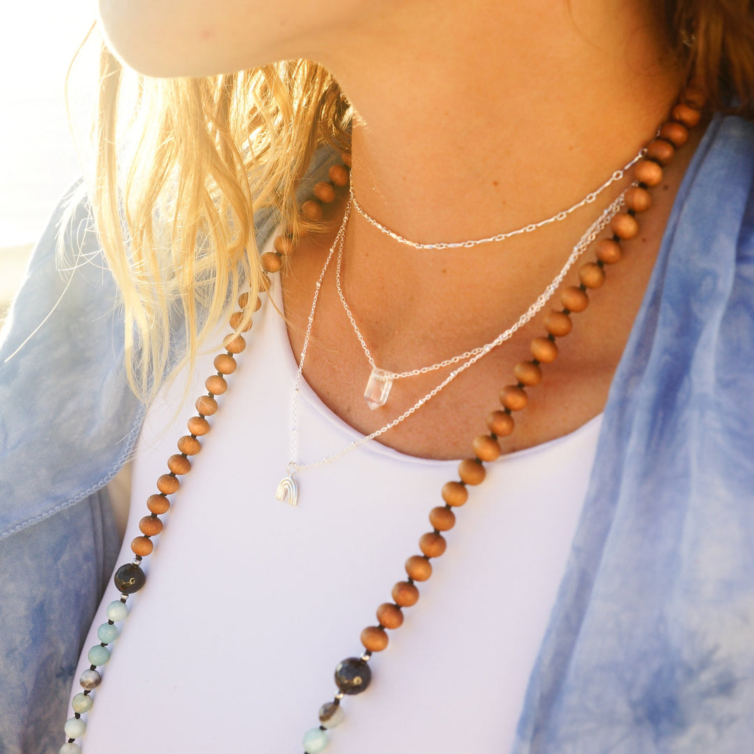 Woman wearing layered necklaces including a clear quartz crystal point necklace, a sterling silver rainbow charm necklace, and Twinkle choker, styled with a beaded mala necklace — boho ocean-inspired jewelry by Blooming Lotus Jewelry.