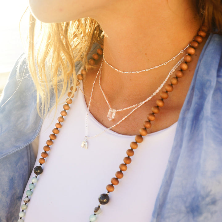Woman wearing layered necklaces including a clear quartz crystal point necklace, a sterling silver rainbow charm necklace, and Twinkle choker, styled with a beaded mala necklace — boho ocean-inspired jewelry by Blooming Lotus Jewelry.