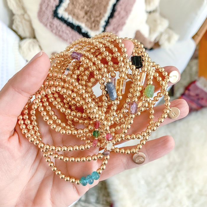 Gold Beaded Bracelets in hand with boho pillows in background - initial disc zodiac gemstones - Blooming Lotus Jewelry