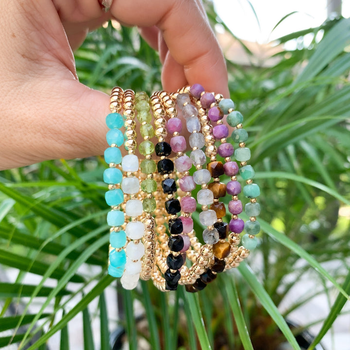 Hand holding a stack of gold beaded bracelets accented with faceted 4mm gemstones in vibrant shades of turquoise, green, pink, black, and gold. Set against a lush green palm background, showcasing a colorful and coastal-inspired jewelry collection.
