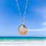 gold shell pendant hanging on gold bead chain with beach ocean in background - blooming lotus jewelry