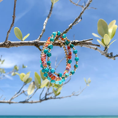 three gold beaded and gemstone bracelets hanging from branch at the beach