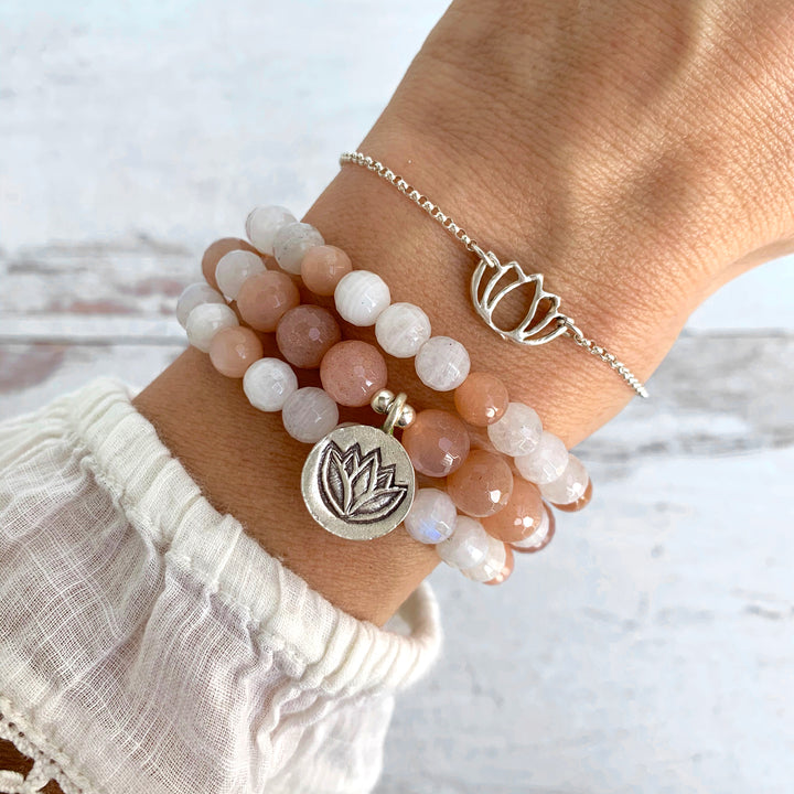 A close-up of a wrist adorned with a yoga jewelry stack of three white and peach moonstone beaded bracelets, each representing new beginnings and emotional balance. The bracelets feature a silver lotus charm, symbolizing spiritual growth and transformation. Also shown is a delicate silver lotus bracelet, adding to the yoga-inspired style. The background is light and airy, emphasizing the mindful and natural beauty of this yoga jewelry set by Blooming Lotus Jewelry.