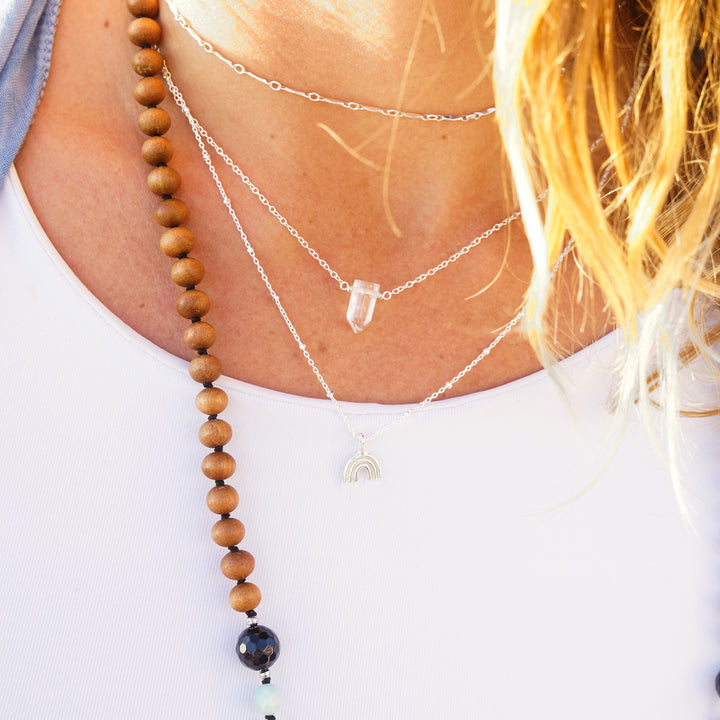 Woman wearing layered necklaces including a clear quartz crystal point necklace, a sterling silver rainbow charm necklace, and Twinkle choker, styled with a beaded mala necklace — boho ocean-inspired jewelry by Blooming Lotus Jewelry.