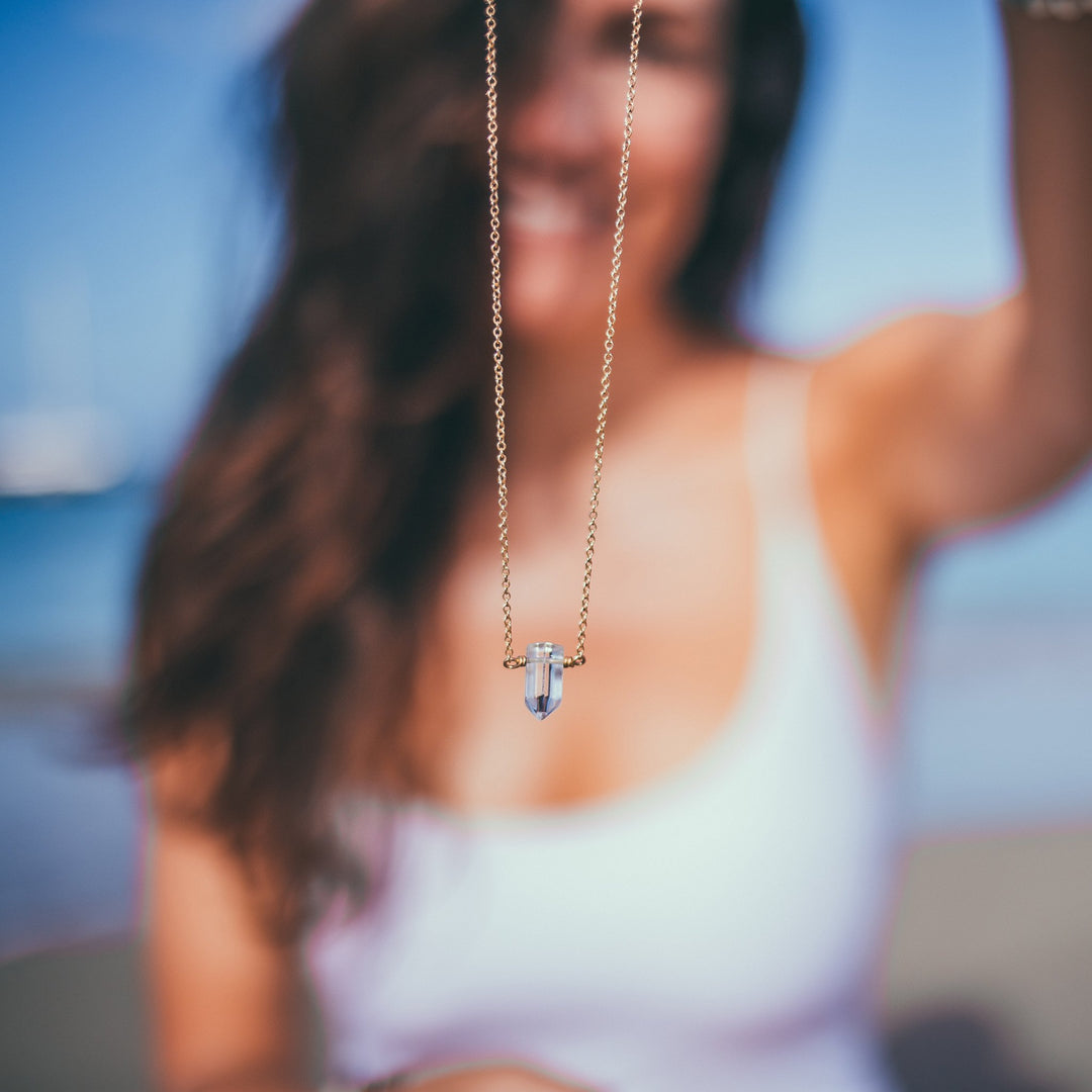 Woman holding a clear quartz crystal point necklace with gold-filled chain in focus, blurred ocean background — yoga-inspired healing crystal jewelry by Blooming Lotus Jewelry.