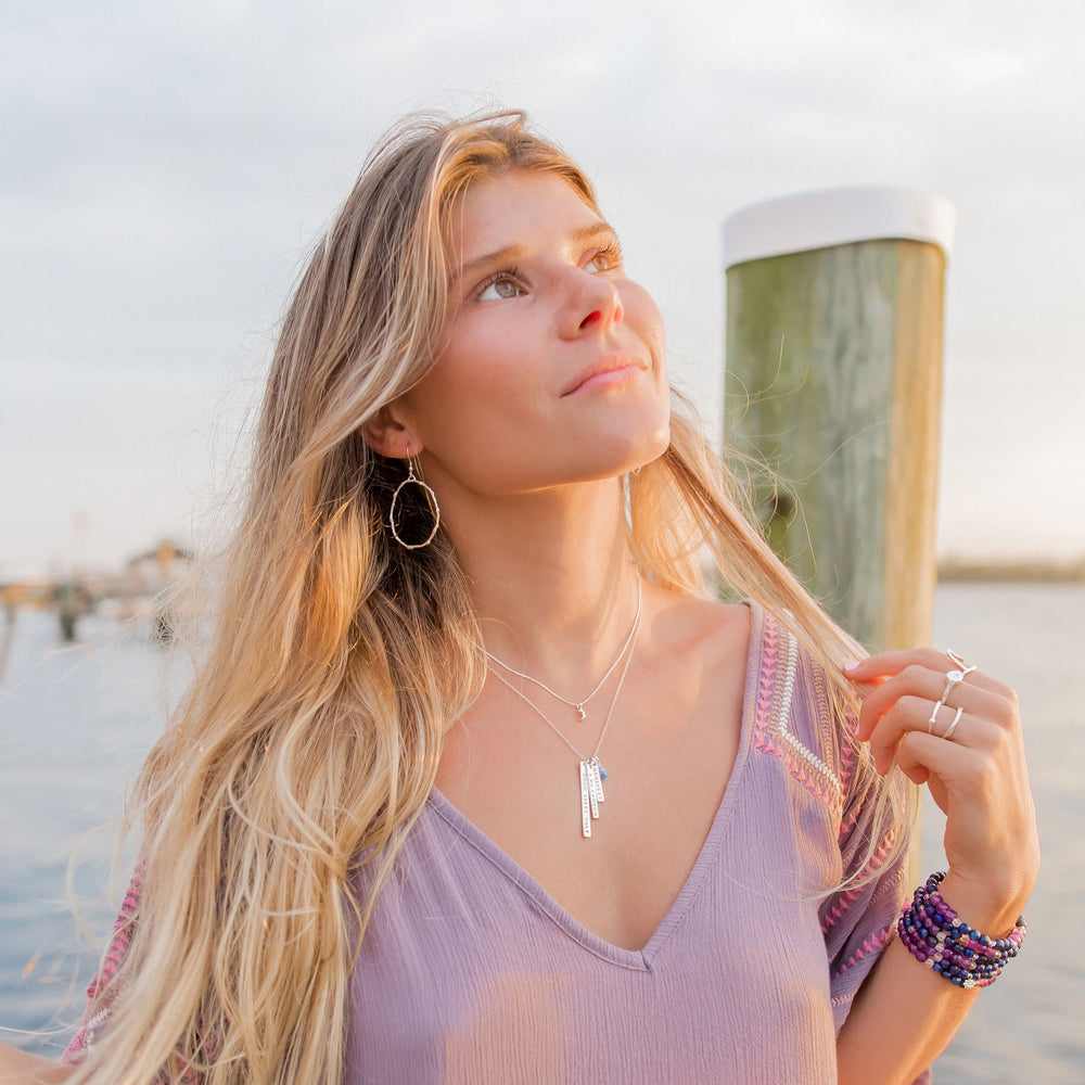 Boho woman wearing layered sterling silver necklaces, including vertical mantra bars with blue gemstone charms and a tiny crescent moon, styled with ocean-inspired rings and gemstone bracelets at golden hour — handmade jewelry by Blooming Lotus Jewelry.
