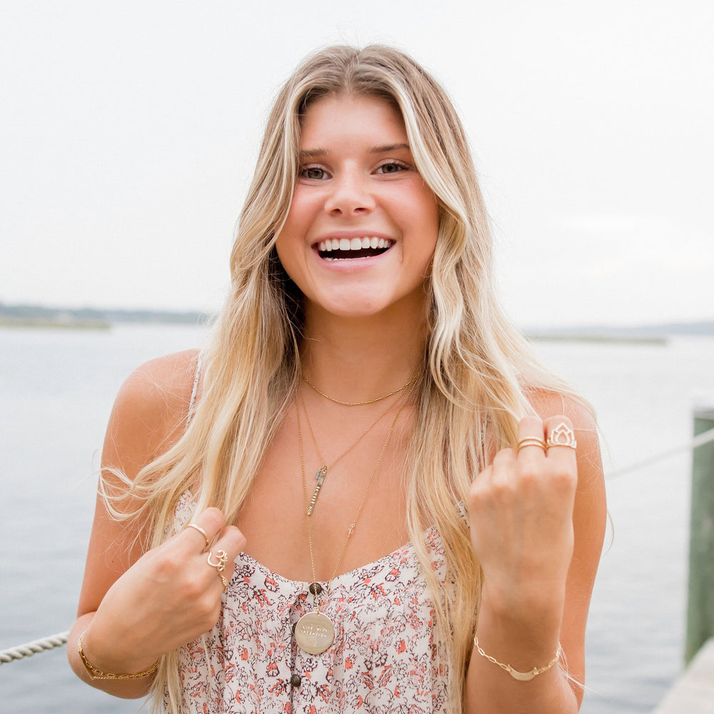 Smiling woman wearing layered gold necklaces including a personalized mantra bar, large coin pendant, and twinkle choker, styled with boho rings and floral dress by the water — handcrafted coastal jewelry by Blooming Lotus Jewelry.